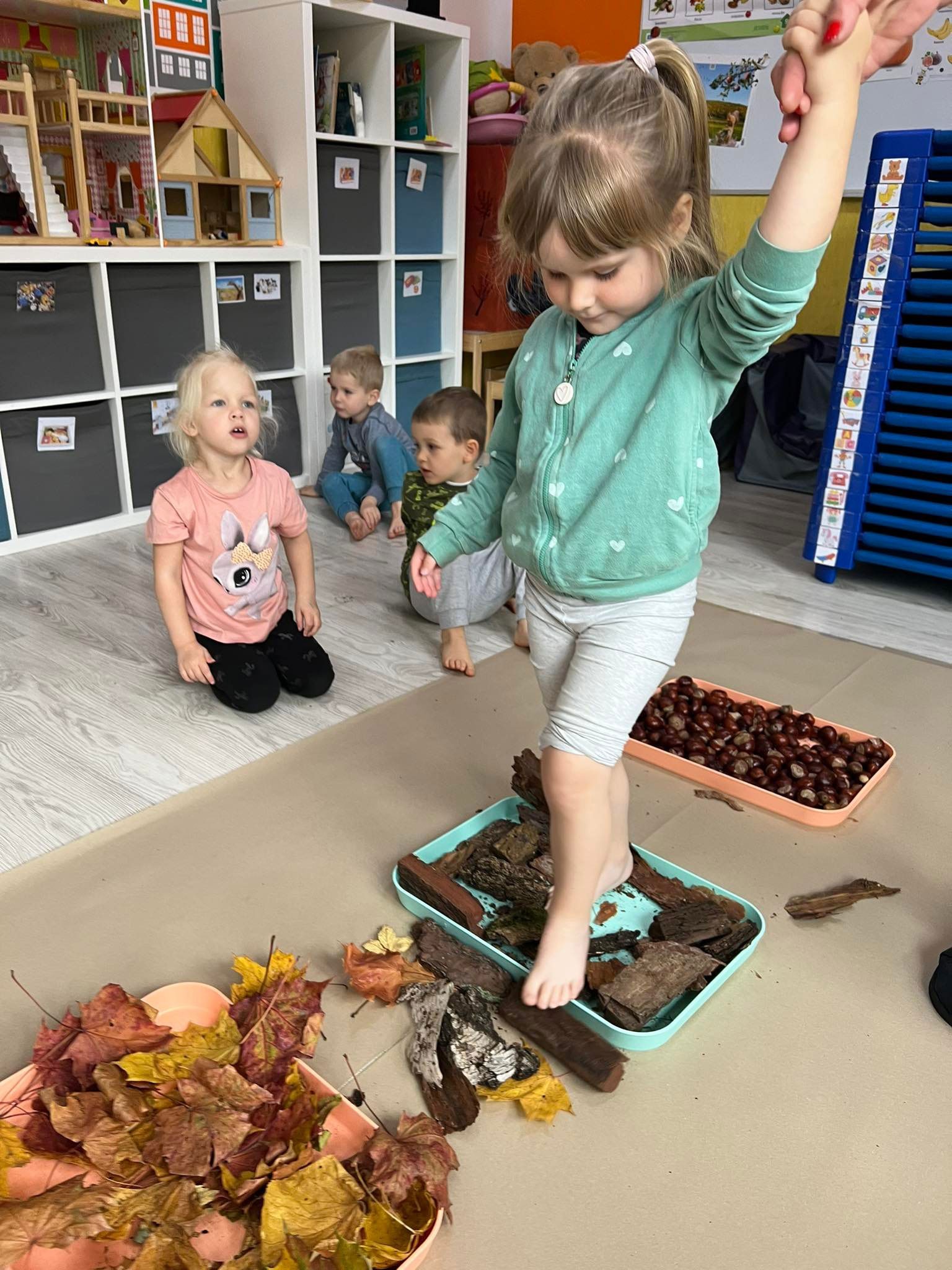 Dziewczynka balansuje stopą na korze drzewa w turkusowej tacy, obok taca z kasztanami i liśćmi, w tle bawiące się dzieci i regał z zabawkami.