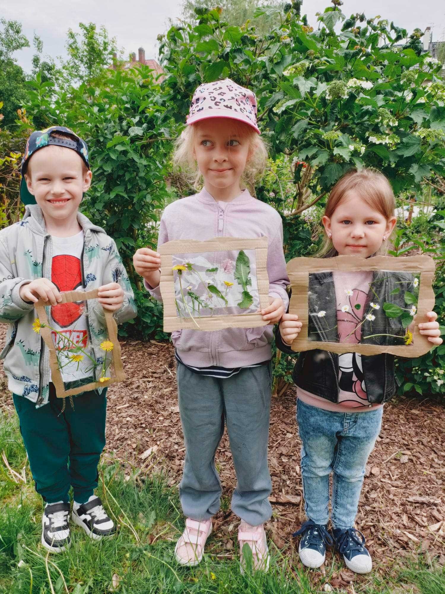 Troje uśmiechniętych dzieci trzyma w rękach ramki z kartonu z wklejonymi kwiatami i liśćmi na tle zieleni.