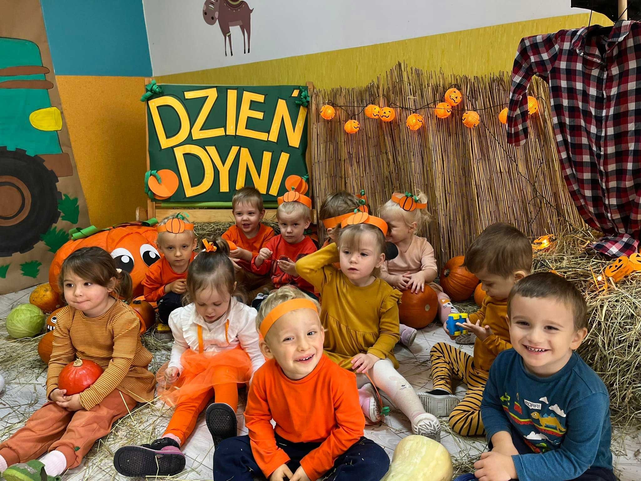 Grupa uśmiechniętych dzieci w pomarańczowych ubraniach i opaskach z dyniami siedzi na sianie na tle dekoracji z okazji Dnia Dyni w żłobku.