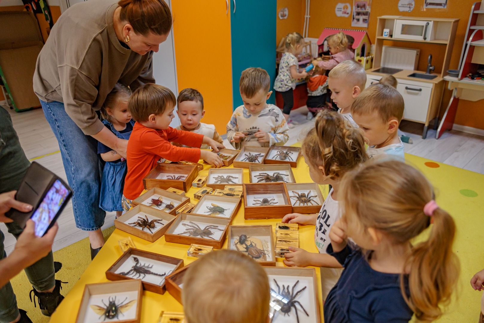 Dzieci z opiekunką przy żółtym stole oglądają eksponaty owadów i pajęczaków w drewnianych gablotkach, w tle widoczna kuchenka zabawkowa.
