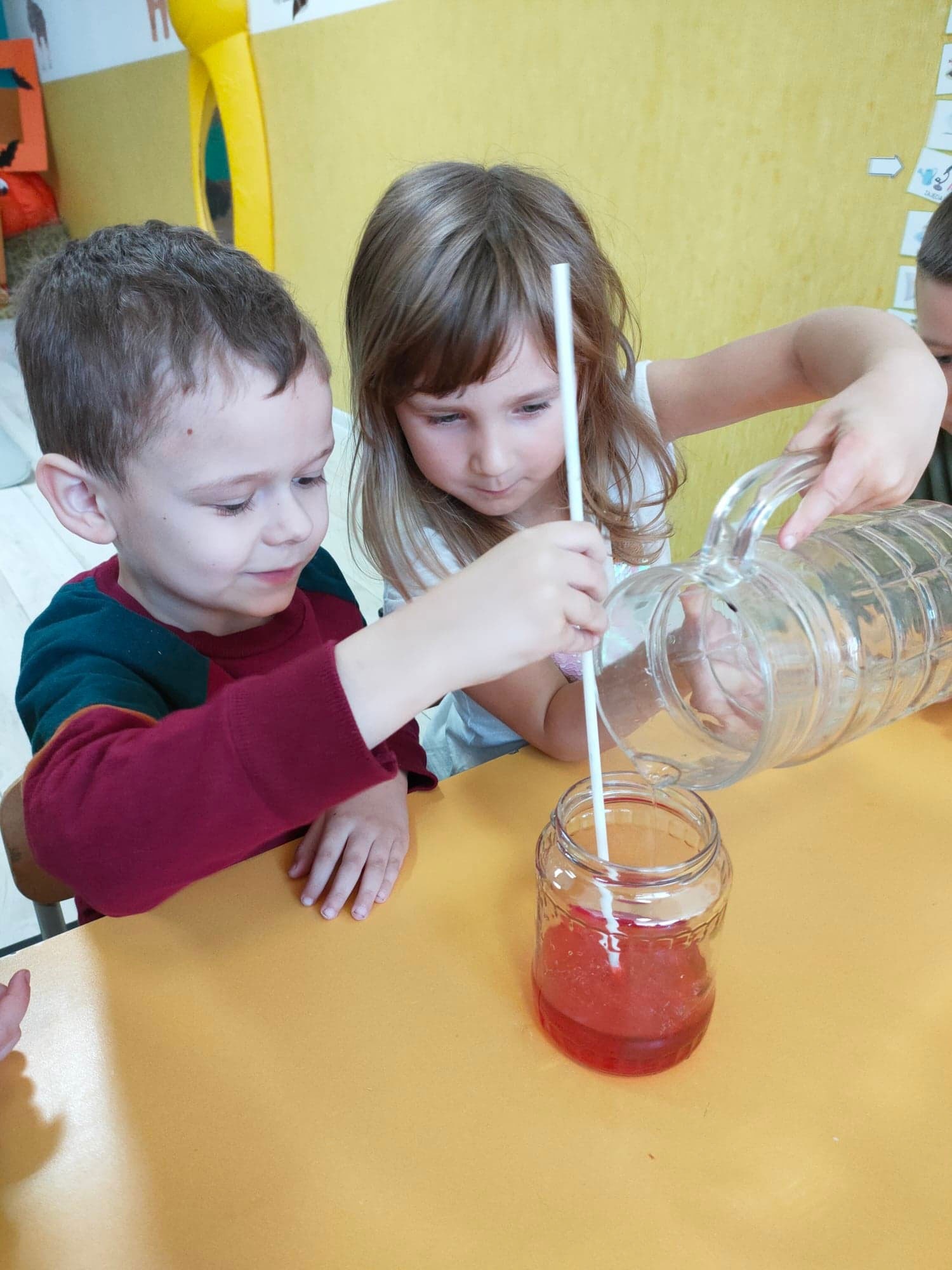 Dwoje dzieci przy żółtym stoliku w trakcie eksperymentu z przelewaniem czerwonego płynu z dzbanka do słoika, w którym znajduje się słomka.