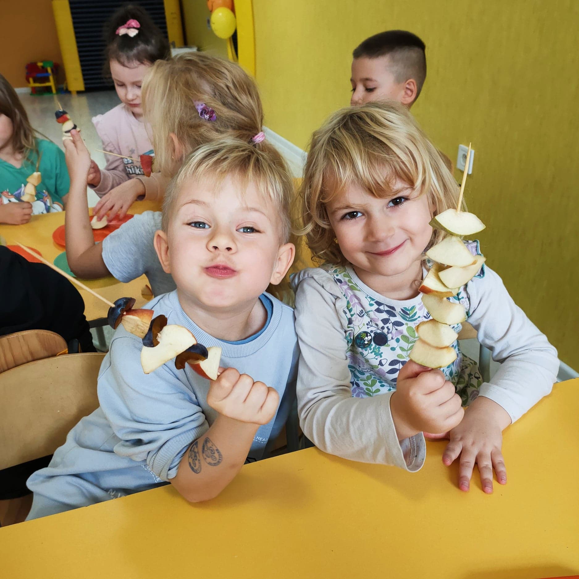 Dwoje uśmiechniętych dzieci trzymających szaszłyki owocowe z jabłek i śliwek przy żółtym stole, widoczne również inne dzieci w tle, na ramieniu chłopca widoczny tatuaż.