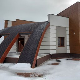 Baronin Development - Nowoczesny dom jednorodzinny z nietypowym dachem pokrytym gontem bitumicznym, elewacja w kolorach bieli, szarości i drewna, zimowy krajobraz z resztkami śniegu.