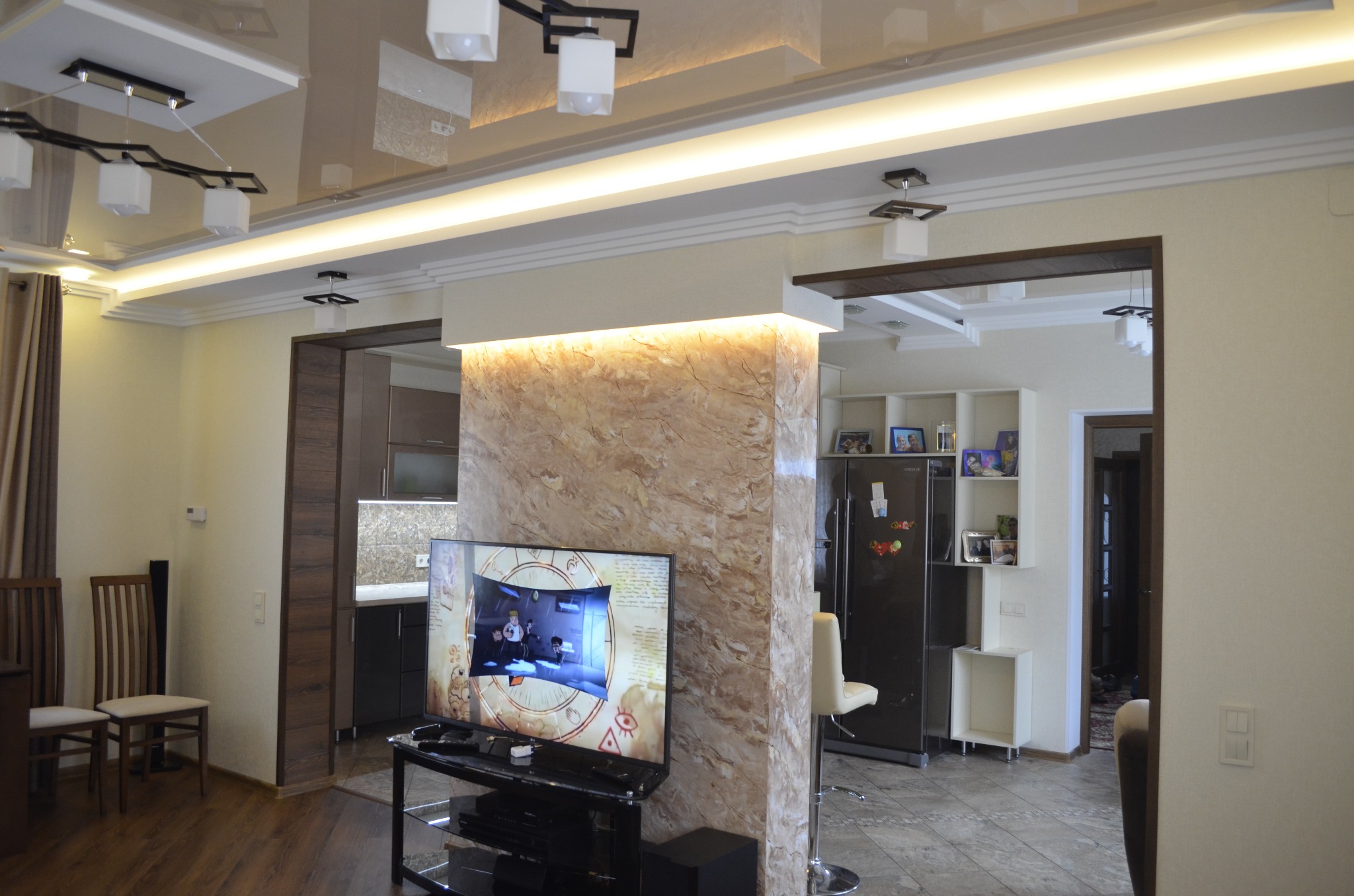 Jasne wnętrze salonu z widoczną ścianą i kolumną wykończoną imitacją marmuru, podwieszany sufit z oświetleniem LED, telewizor na stoliku RTV, widok na kuchnię w tle.