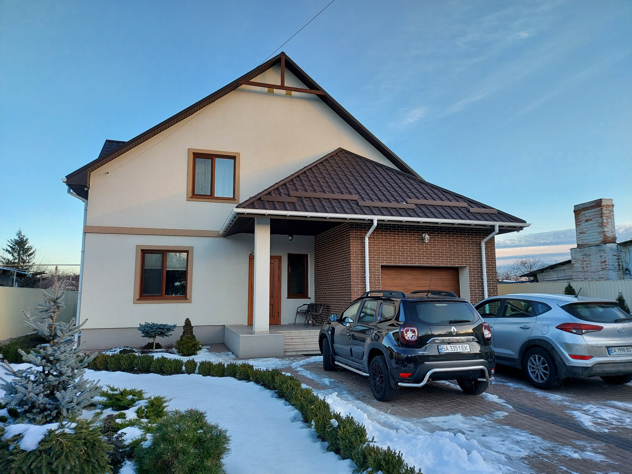 Ocieplony dom jednorodzinny z elewacją w kolorze kremowym i brązowym dachem, z garażem obłożonym czerwoną cegłą klinkierową, widok z zewnątrz w zimowej scenerii z dwoma zaparkowanymi samochodami.