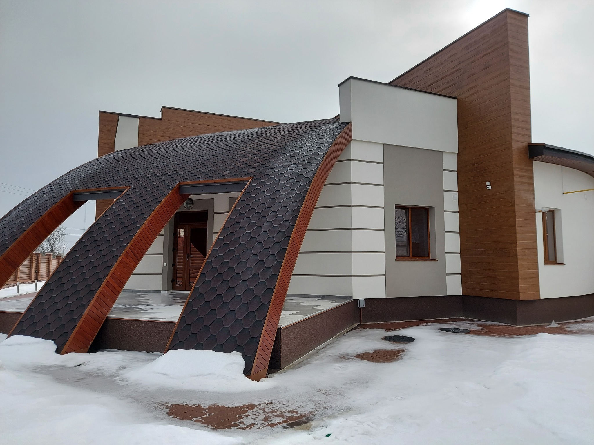 Nowoczesny dom jednorodzinny z nietypowym dachem pokrytym gontem bitumicznym, elewacja w kolorach bieli, szarości i drewna, zimowy krajobraz z resztkami śniegu.