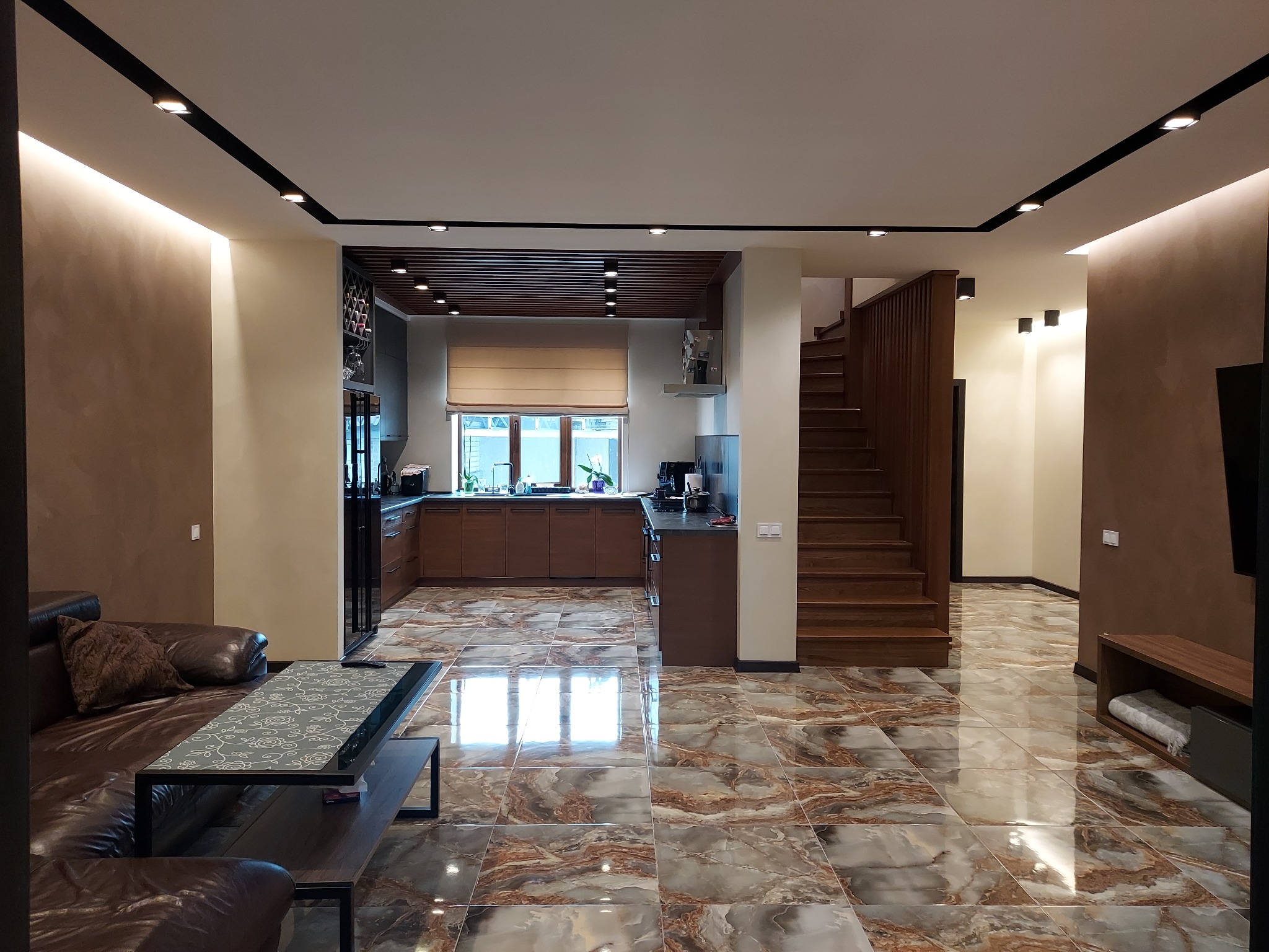 Przestronne wnętrze domu z marmurową podłogą, drewnianymi schodami i kuchnią w tle, oświetlone punktowymi lampami sufitowymi.