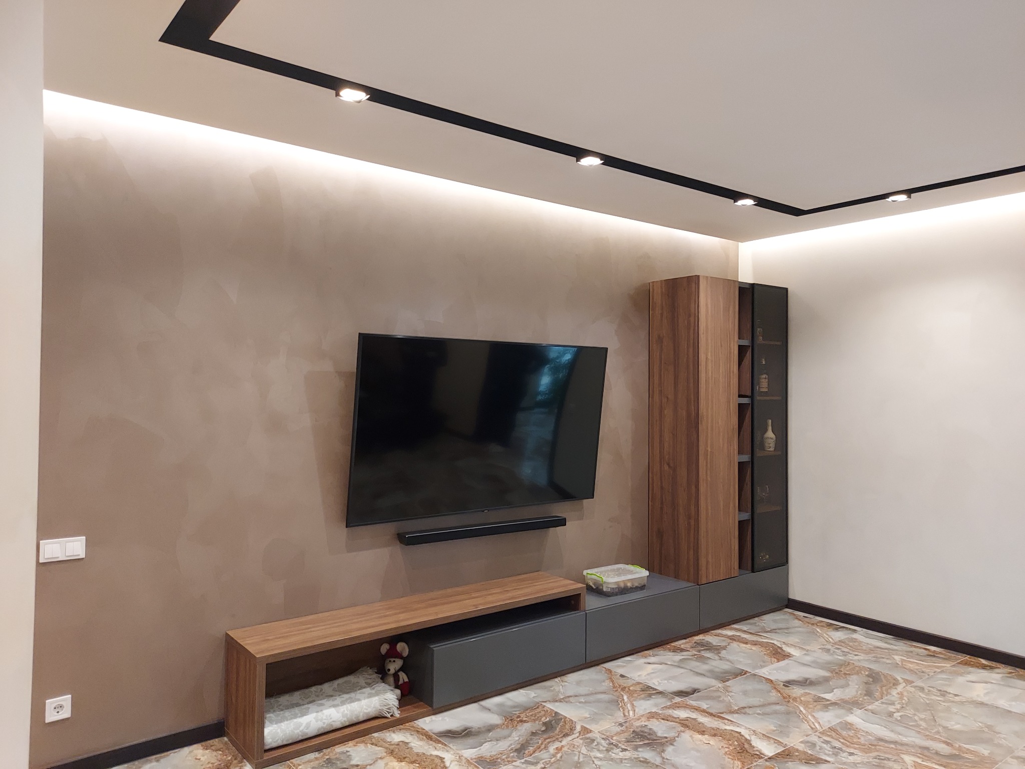 Nowoczesny salon z telewizorem na ścianie o beżowej, dekoracyjnej fakturze, oświetleniem punktowym na suficie i meblami w odcieniach drewna i szarości, wykończony płytkami podłogowymi imitującymi...