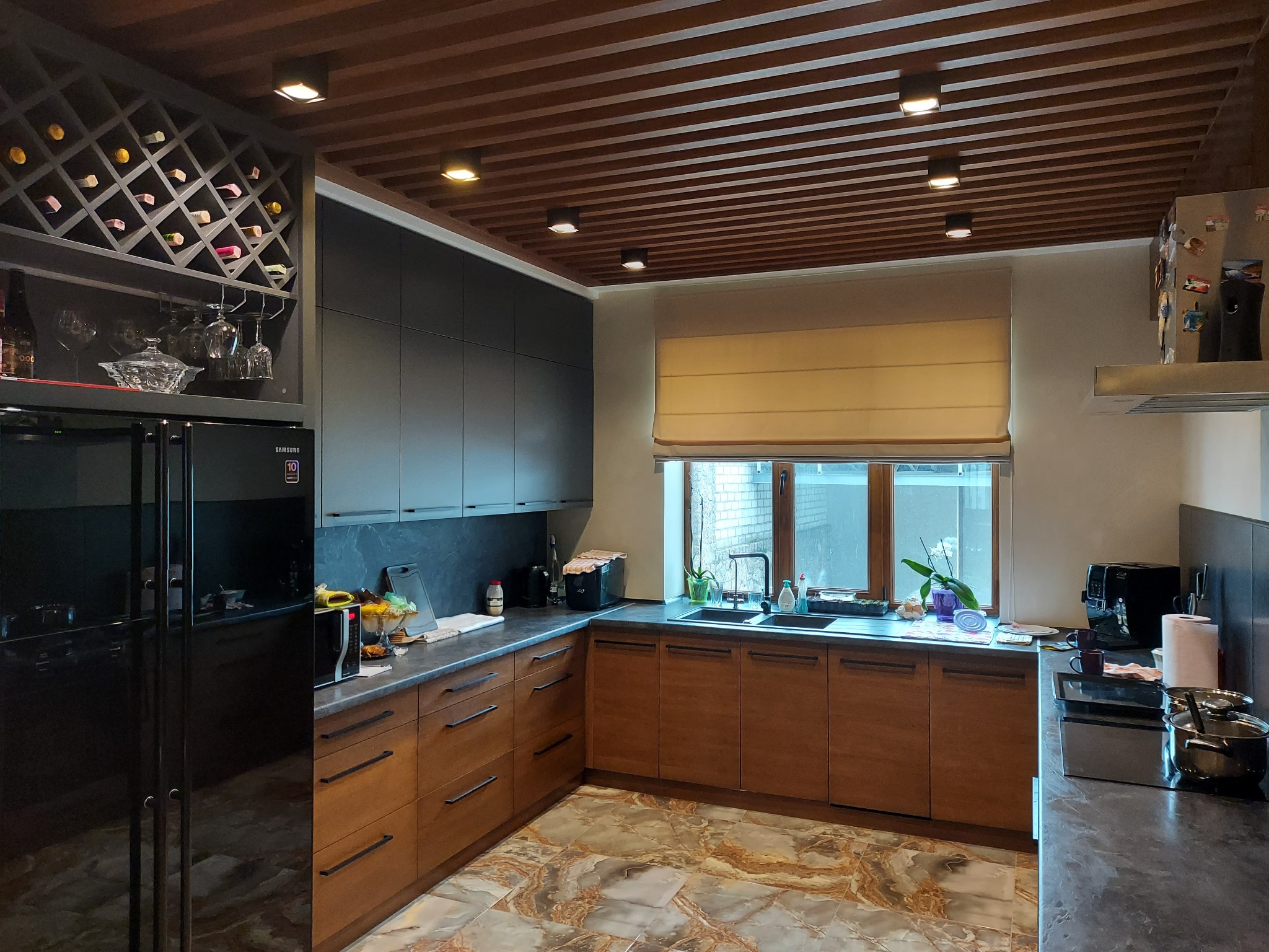 Nowoczesna kuchnia z drewnianymi szafkami dolnymi i grafitowymi górnymi, marmurową podłogą, drewnianym sufitem lamelowym i wbudowaną chłodziarką do wina.