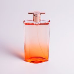 Pure Mateusz Pisarski - Butelka perfum Lancôme Idôle z gradientem od pomarańczowego do przezroczystego, na białym tle, z metalowym atomizerem w kolorze różowego złota.