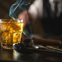 Pure Mateusz Pisarski - Szklanka whisky z lodem, dymiące cygaro i szczypce do lodu na ciemnym blacie, w tle zamazana sylwetka osoby w białej koszuli i czarnej kamizelce.