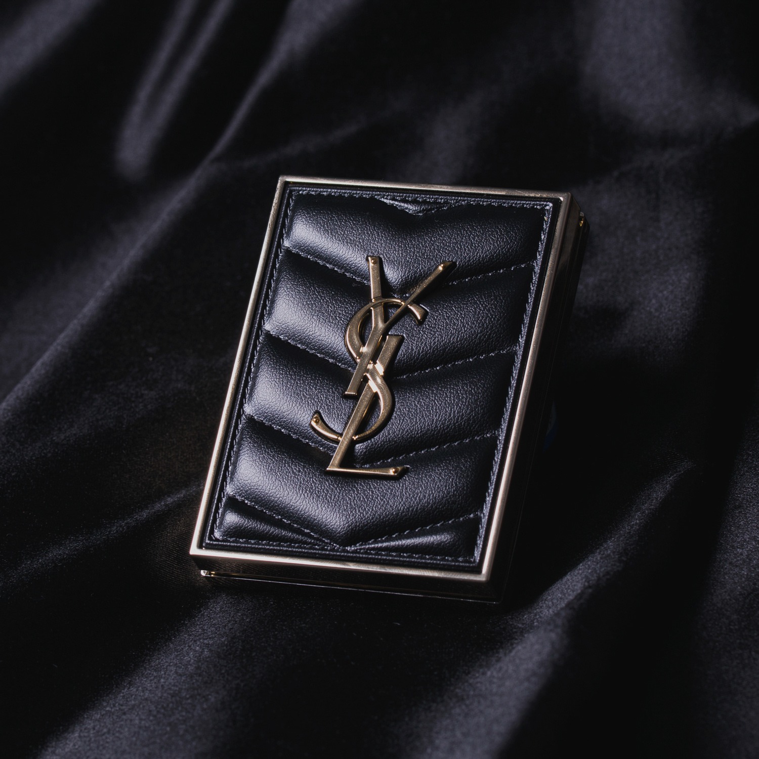 Czarny, pikowany portfel z metalowym logo YSL na tle czarnej, satynowej tkaniny.