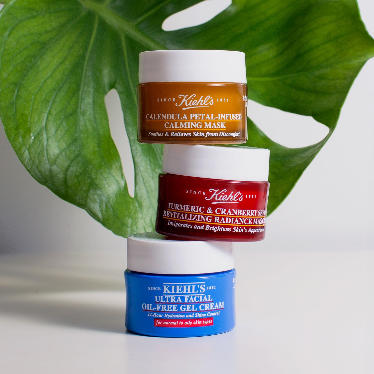 Stos trzech słoiczków z kosmetykami marki Kiehl's na białym tle z zielonym liściem monstery w tle. Od góry: pomarańczowa maska Calendula, czerwona maska Turmeric & Cranberry Seed, niebieski żel...