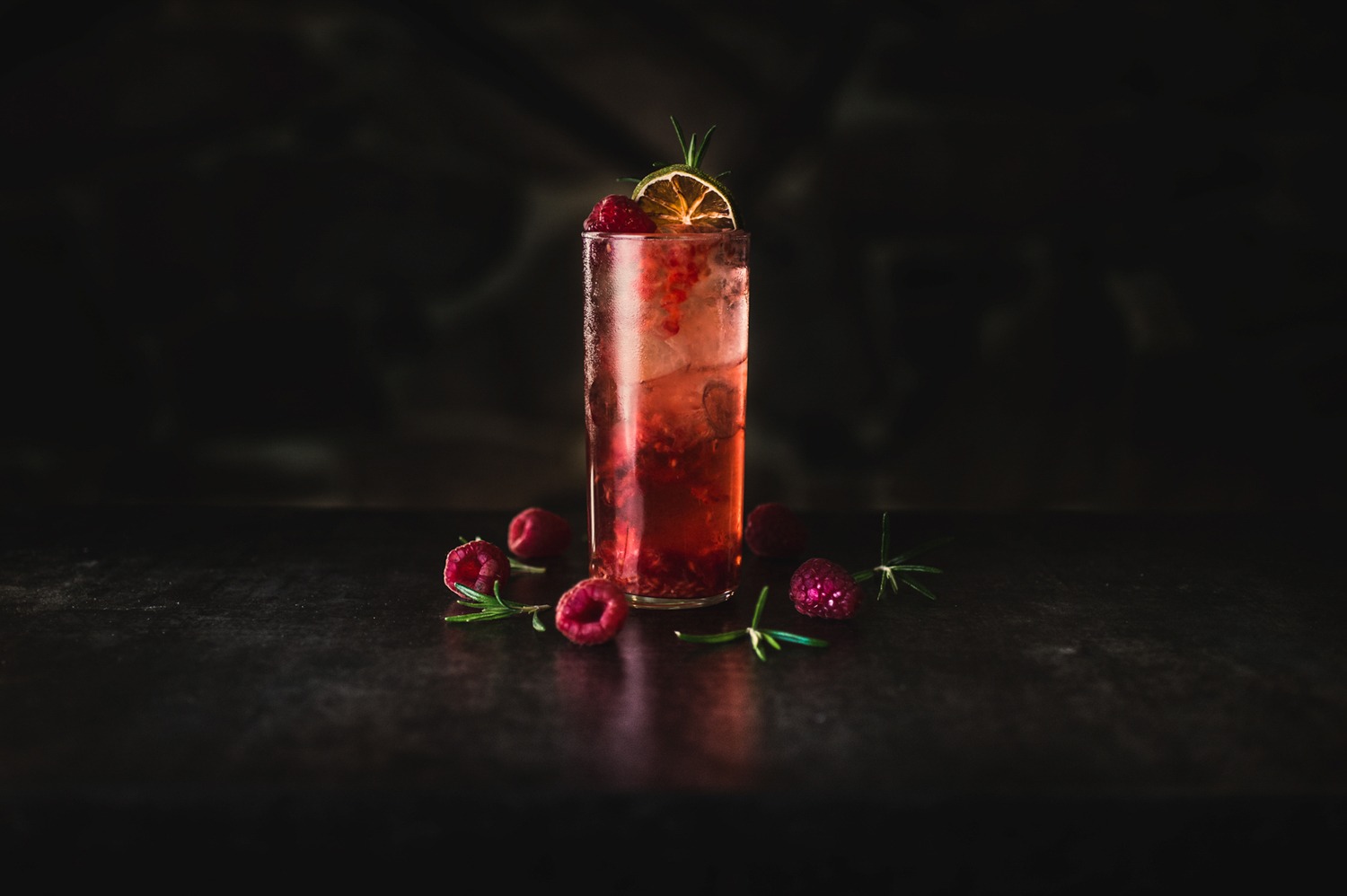 Wysoki drink z malinami, lodem i plasterkiem suszonej limonki na ciemnym tle.