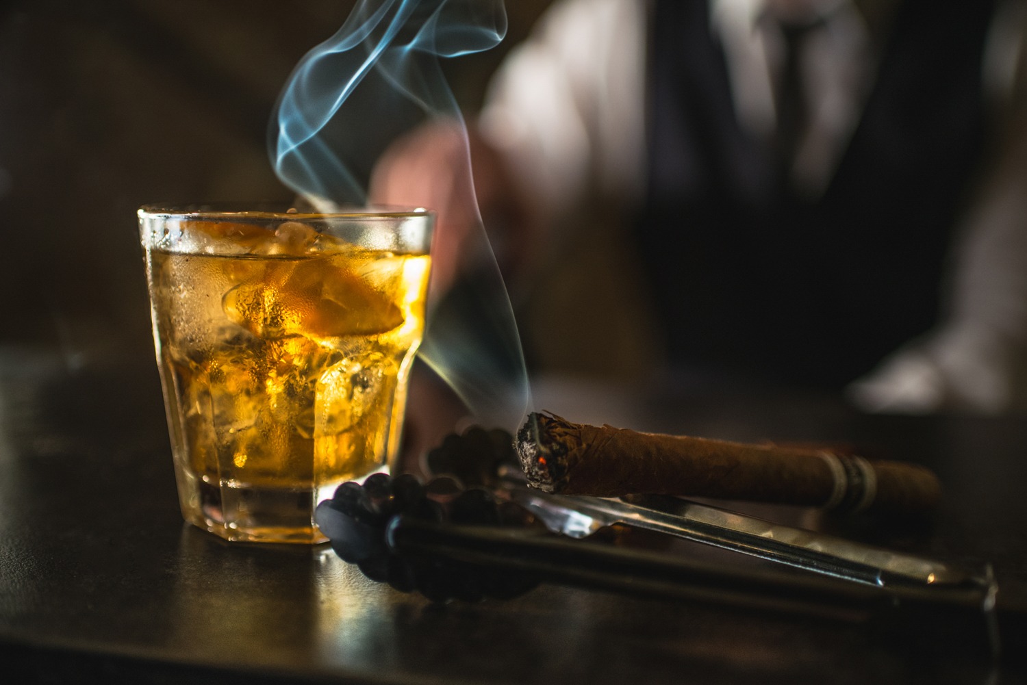 Szklanka whisky z lodem, dymiące cygaro i szczypce do lodu na ciemnym blacie, w tle zamazana sylwetka osoby w białej koszuli i czarnej kamizelce.