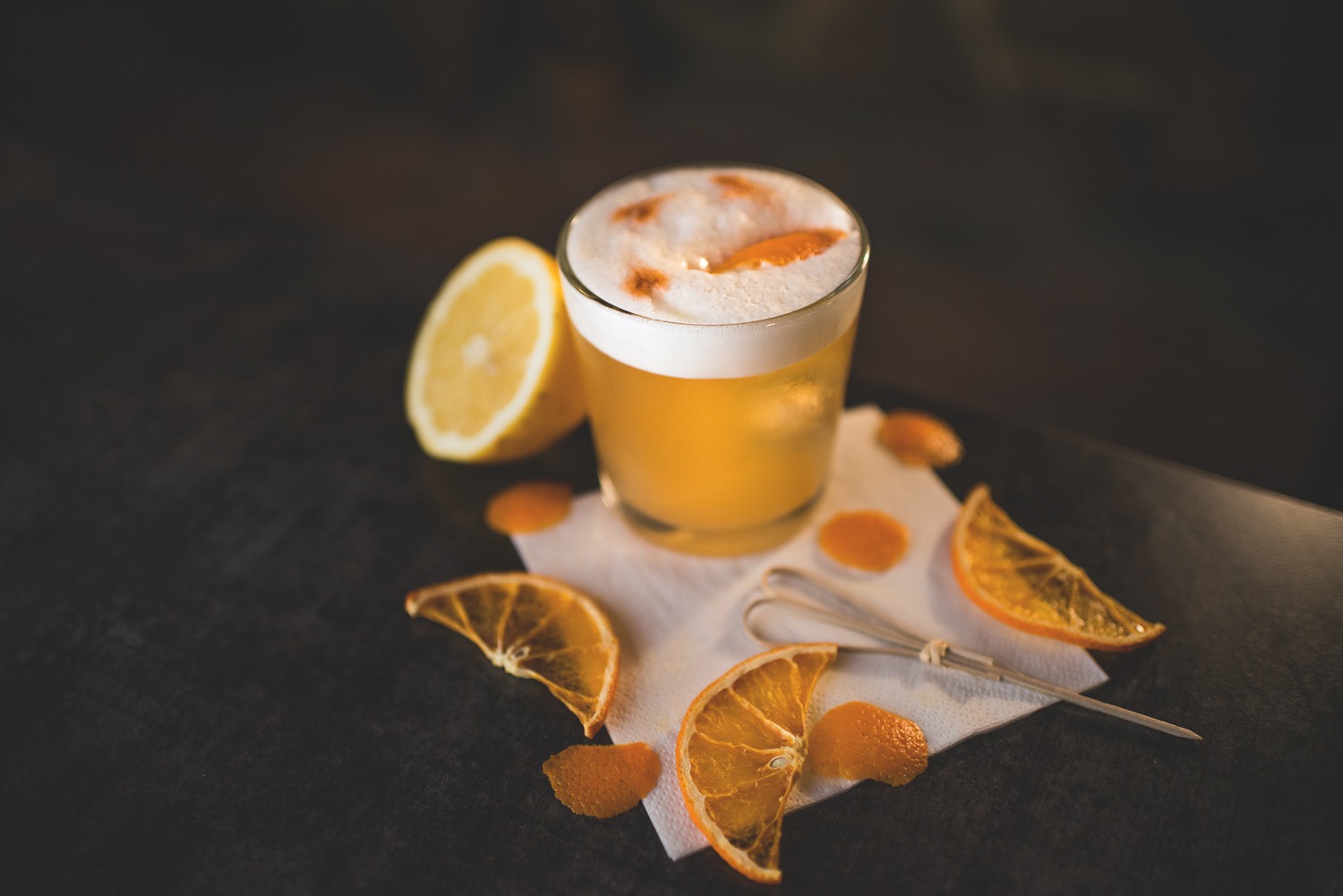 Złocisty koktajl z pianką w niskiej szklance, ozdobiony skórką pomarańczową, obok połówka cytryny, suszone plastry pomarańczy i mieszadełko na białej serwetce na ciemnym blacie.
