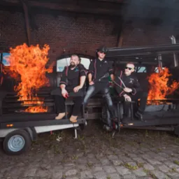 Trzech kucharzy w czarnych uniformach siedzi na przyczepie z grillem, z którego wydobywają się płomienie, na tle ceglanej ściany.