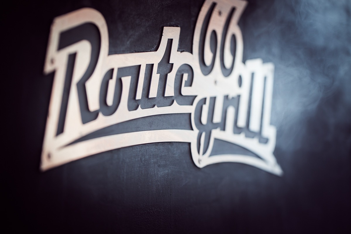 Metalowy szyld 'Route 66 Grill' na ciemnym tle, oświetlony z boku, z delikatną mgłą w tle.