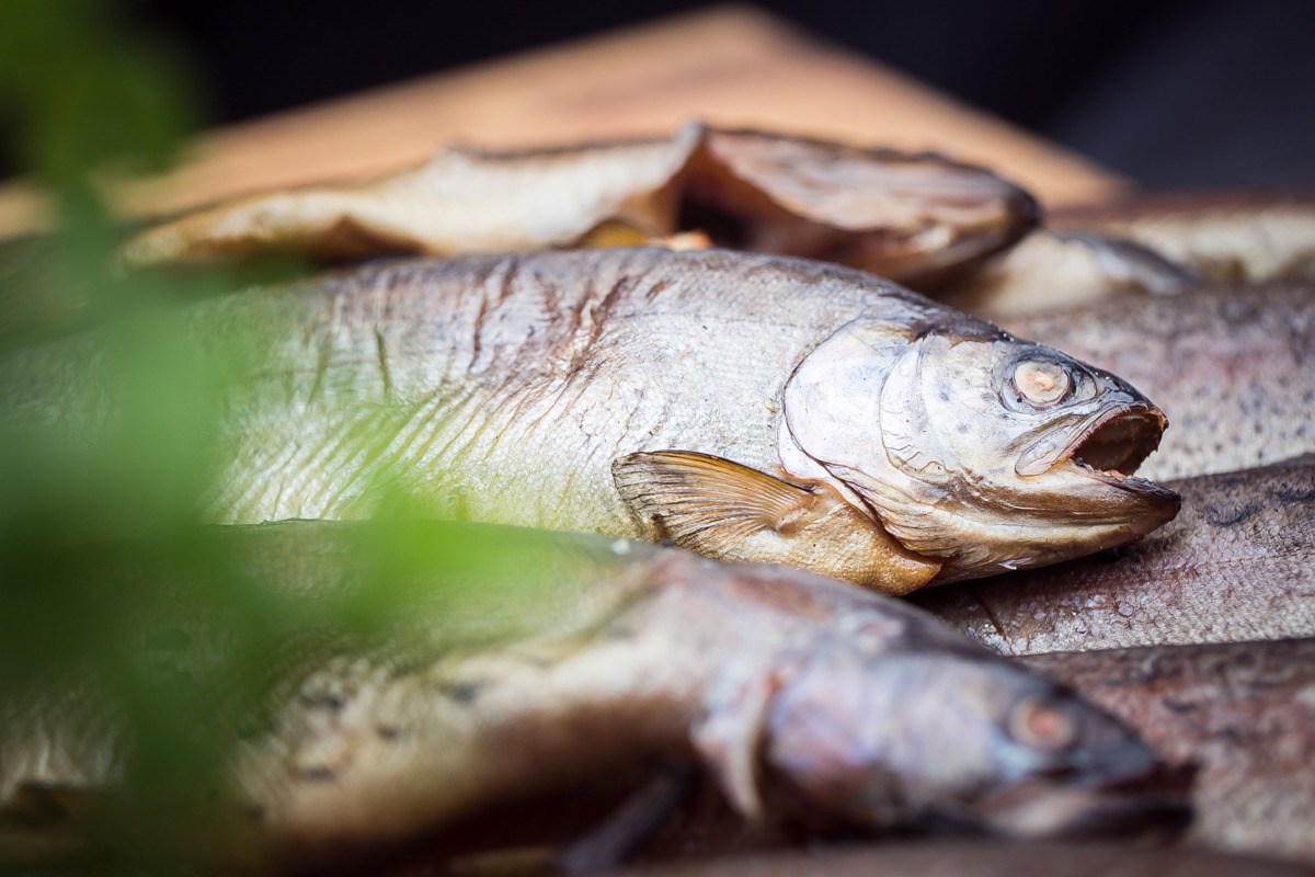 Wędzone pstrągi ułożone na drewnianej desce, z zielonym akcentem roślinnym w lewym dolnym rogu kadru, ukazane w zbliżeniu.
