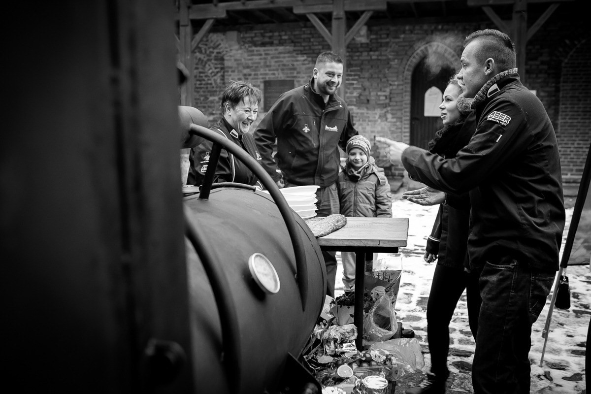 Czarno-białe zdjęcie przedstawia grupę osób w plenerze, prawdopodobnie podczas wydarzenia plenerowego z grillem. W centrum kadru widoczny duży grill z termometrem, obok którego stoi stół...