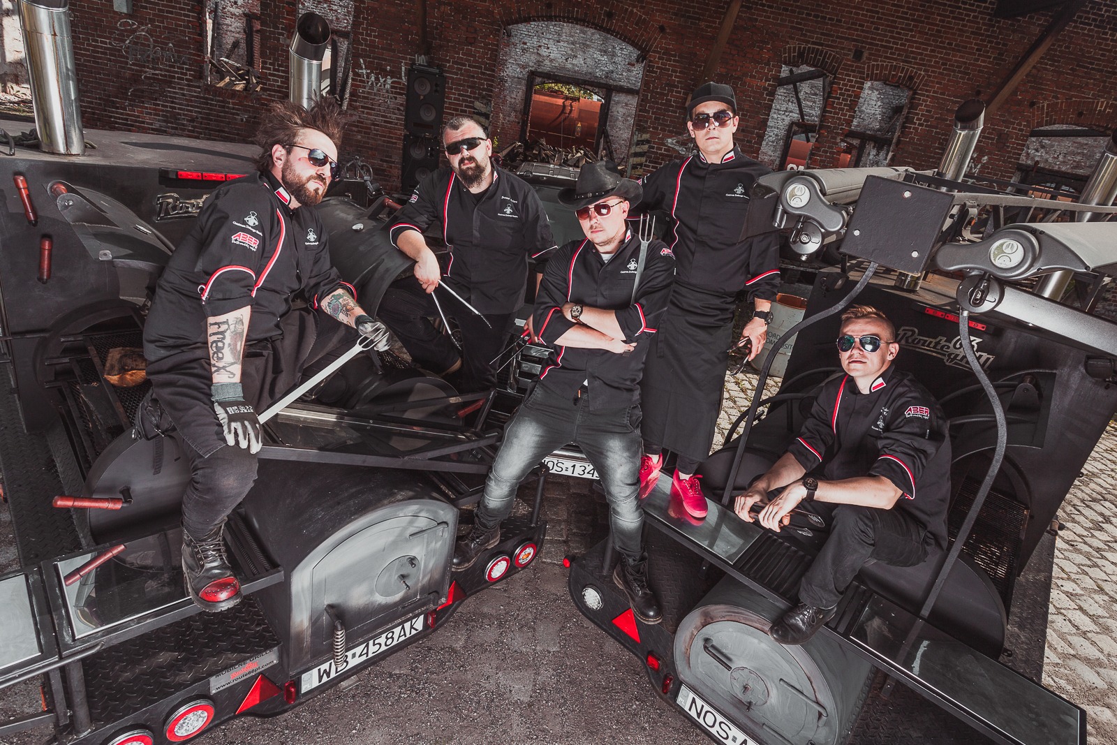 Grupa pięciu kucharzy w czarnych uniformach z czerwonymi detalami i okularach przeciwsłonecznych pozuje na tle mobilnych grilli w industrialnym otoczeniu ceglanego budynku.