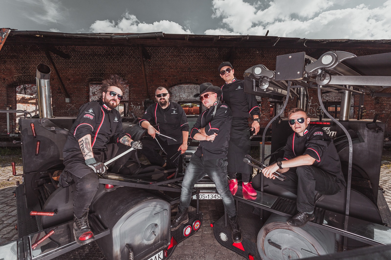 Grupa kucharzy w czarnych uniformach z czerwonymi akcentami pozuje na tle ceglanego budynku, stojąc na mobilnej platformie z grillem, trzymając narzędzia kuchenne.