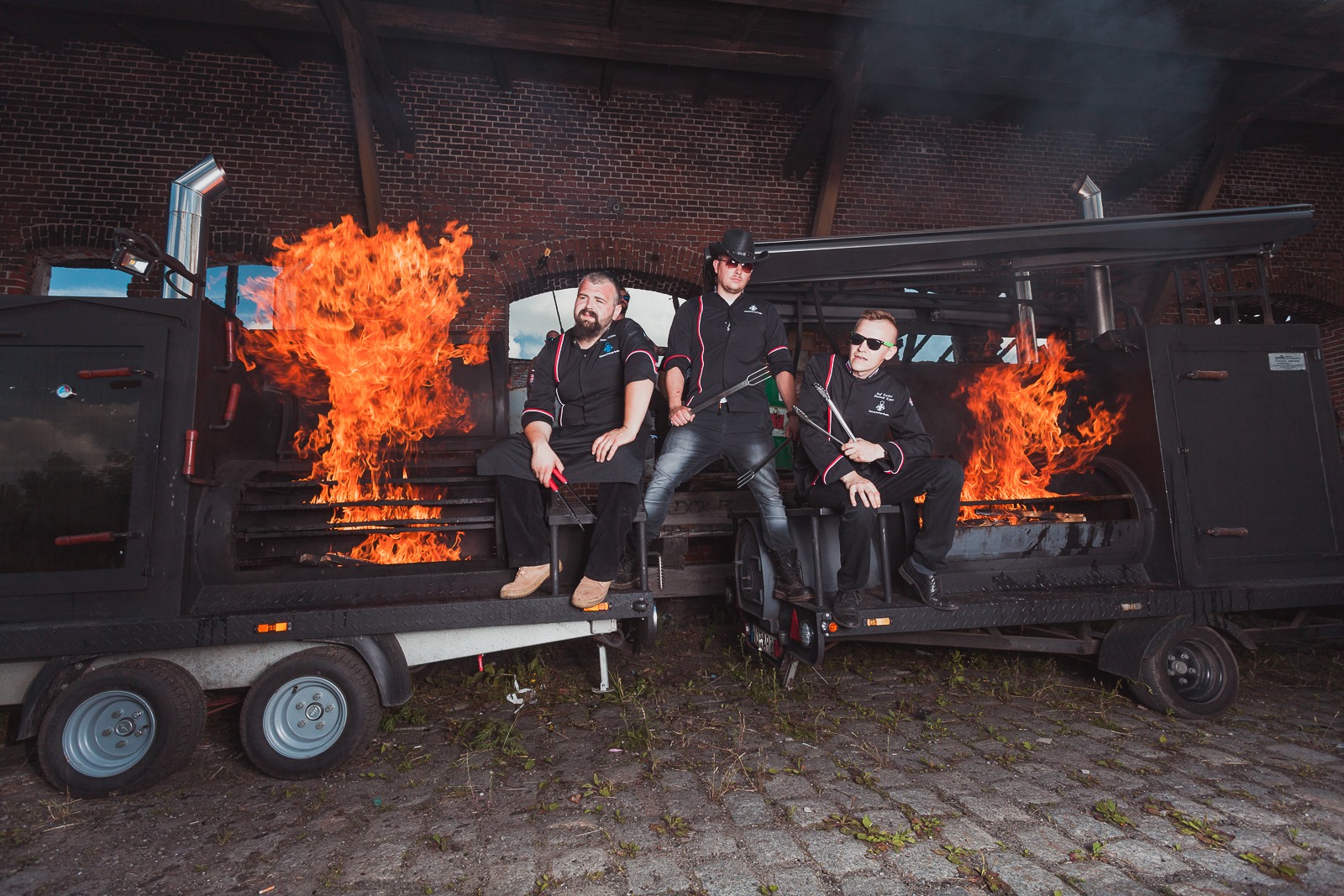Trzech kucharzy w czarnych uniformach siedzi na przyczepie z grillem, z którego wydobywają się płomienie, na tle ceglanej ściany.