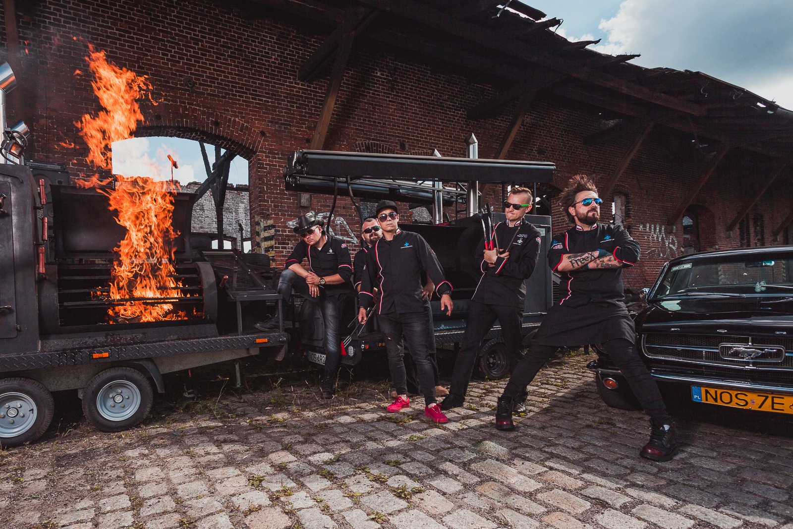 Ekipa kucharzy pozuje na tle rozpalonego grilla na przyczepie i czarnego Mustanga, ceglany budynek w tle.