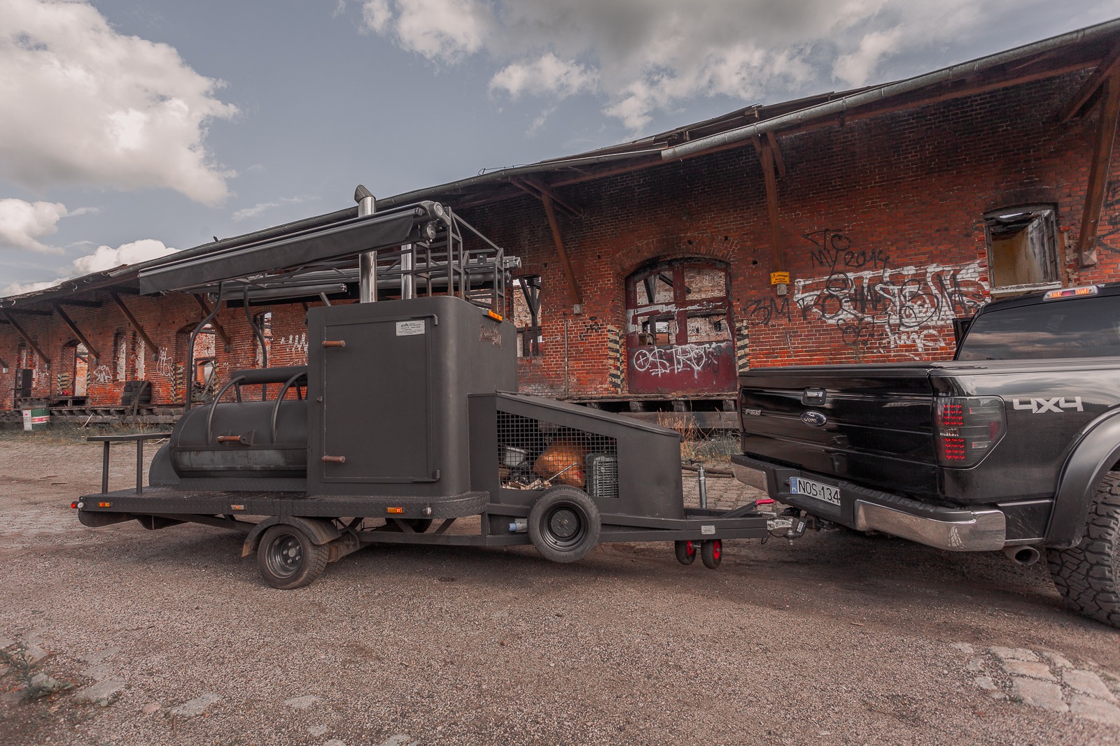 Czarny mobilny grill w formie przyczepy samochodowej, zaparkowany przed ceglanym budynkiem z graffiti, podpięty do czarnego pickupa.