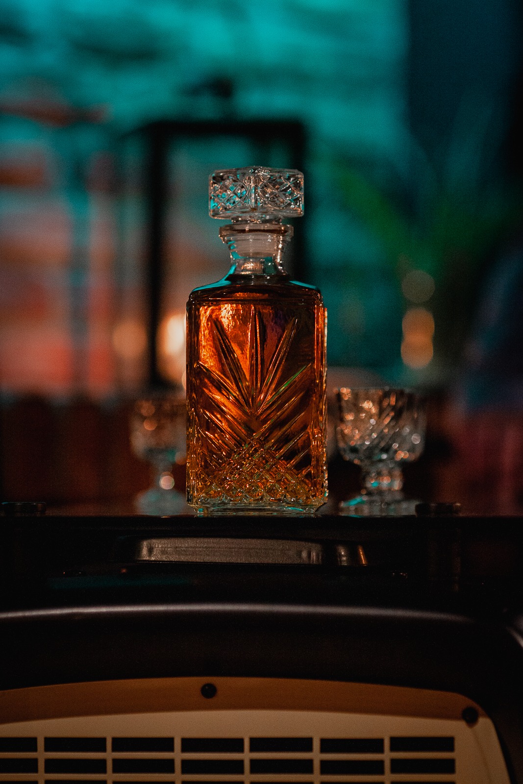 Kryształowa karafka z bursztynowym trunkiem i dwa kieliszki na barowym blacie, podświetlone turkusowym światłem, z widocznymi detalami kryształowego wzoru.