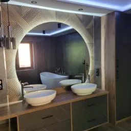 Nowoczesna łazienka z podświetlanym, okrągłym lustrem, dwiema owalnymi umywalkami na drewnianym blacie z szufladami o wzorze marmuru, oraz białą wanną wolnostojącą widoczną w odbiciu lustra.