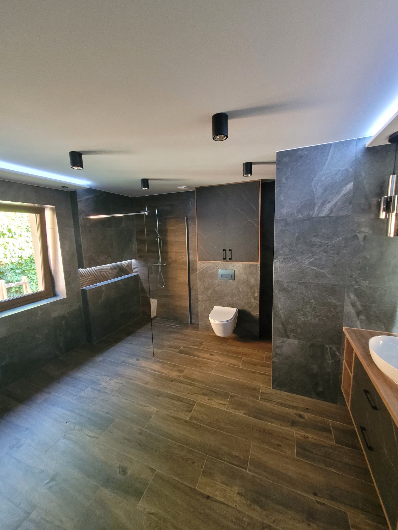 Nowoczesna łazienka z szarymi płytkami imitującymi kamień, drewnopodobną podłogą ułożoną w jodełkę, kabiną prysznicową typu walk-in, podświetleniem LED i wiszącą toaletą.