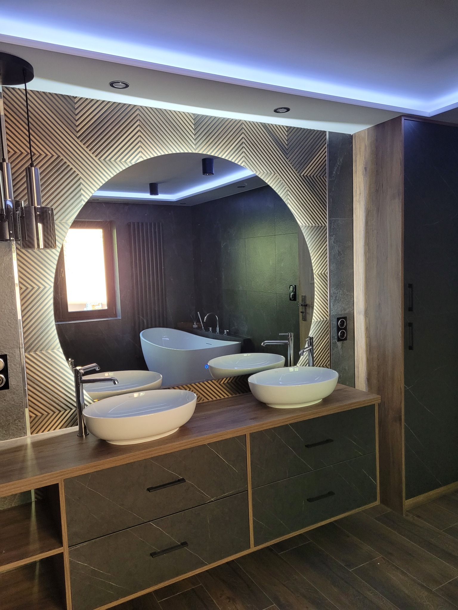 Nowoczesna łazienka z podświetlanym, okrągłym lustrem, dwiema owalnymi umywalkami na drewnianym blacie z szufladami o wzorze marmuru, oraz białą wanną wolnostojącą widoczną w odbiciu lustra.