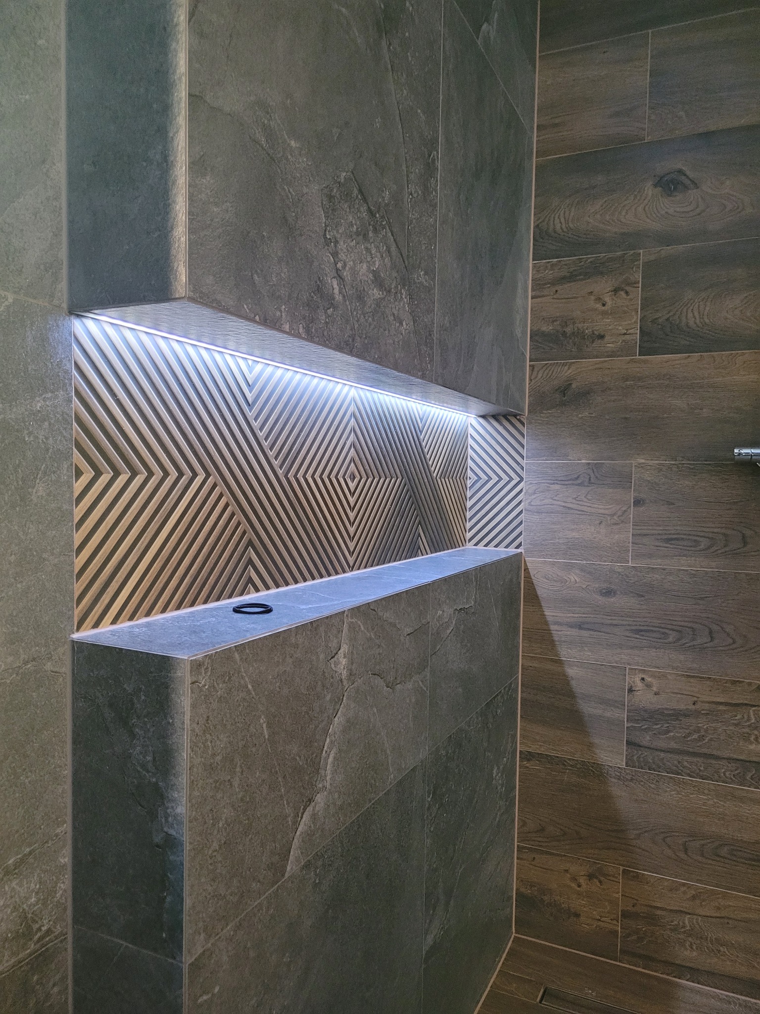 Nowoczesna łazienka z półką wnękową podświetloną LED, wykończona płytkami imitującymi kamień i drewno, z czarnym pierścieniem leżącym na półce.