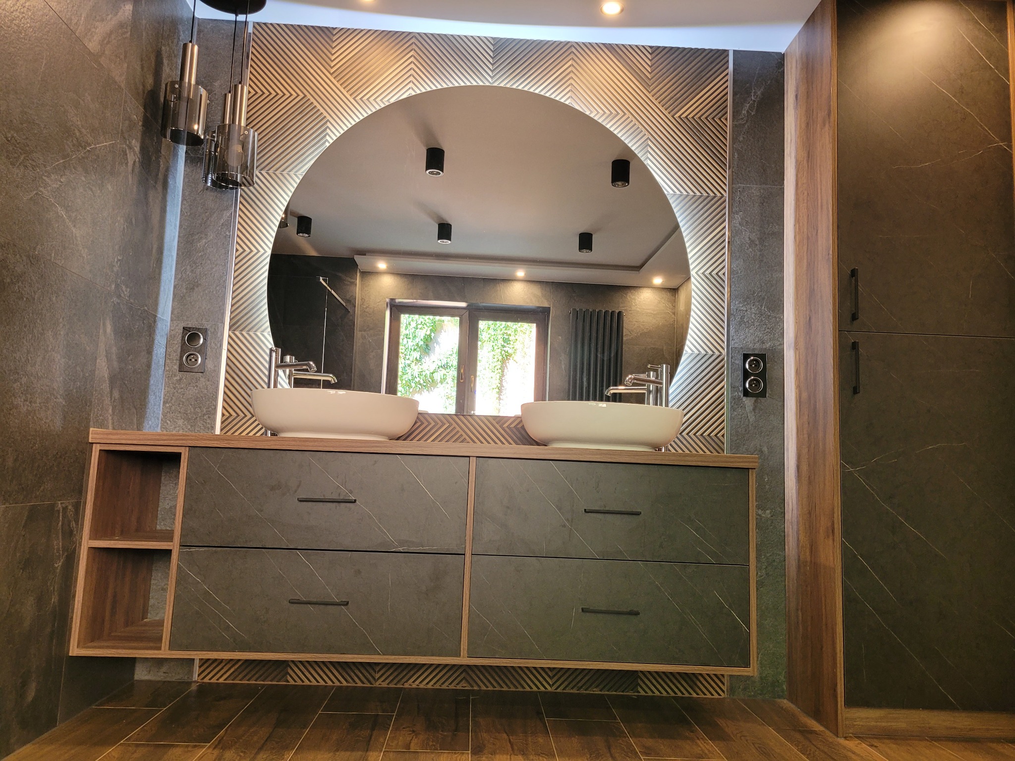 Nowoczesna łazienka z podwójną umywalką nablatową, szafką z szufladami o fakturze kamienia, okrągłym lustrem z podświetleniem i dekoracyjną ścianą z geometrycznym wzorem.