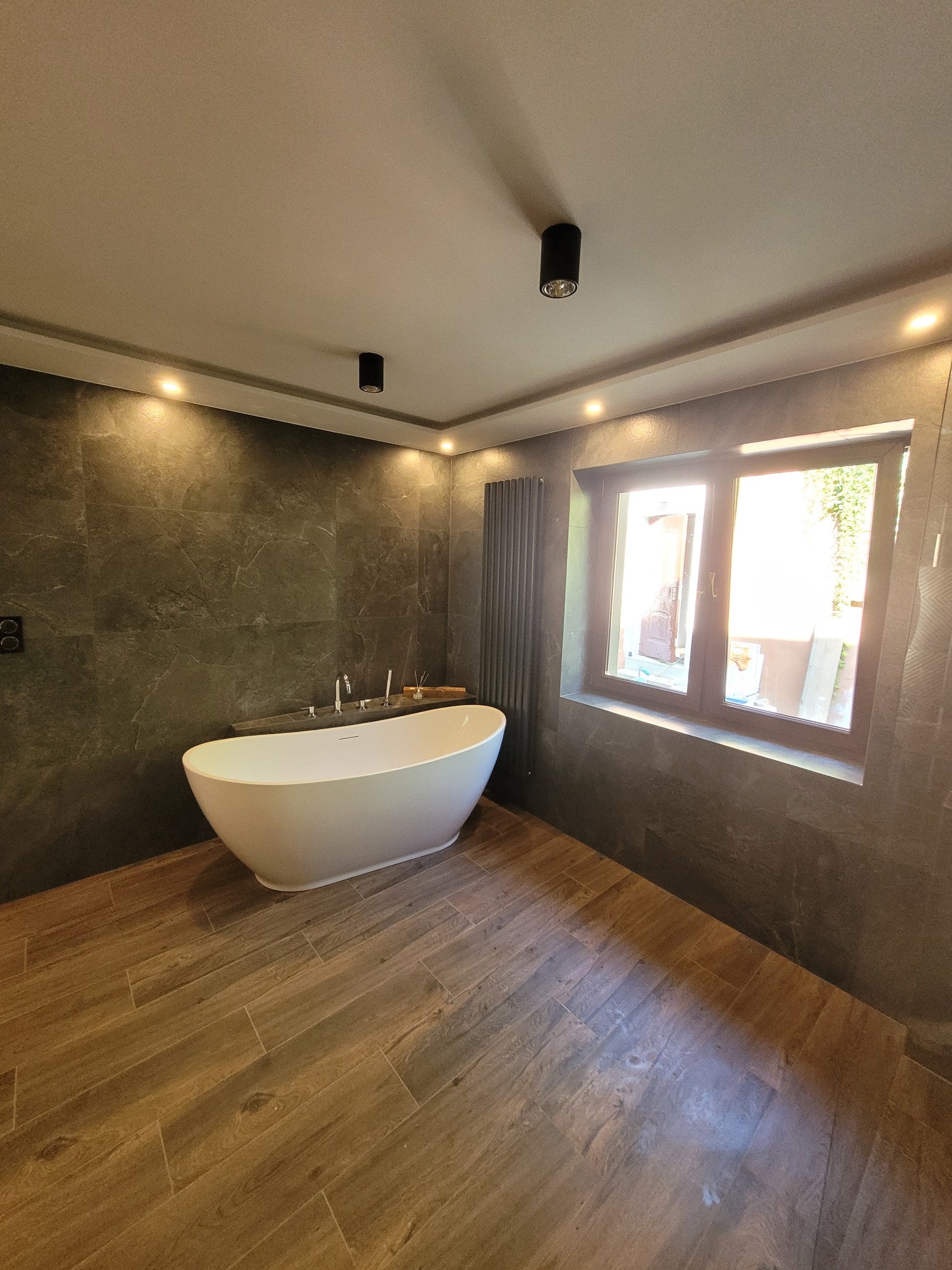 Nowoczesna łazienka z wolnostojącą wanną, szarymi płytkami imitującymi kamień na ścianach, drewnopodobną podłogą, grzejnikiem drabinkowym i oknem z widokiem na zieleń.