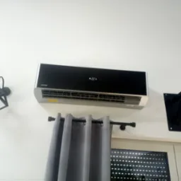 Nowoczesny klimatyzator Sinclair zamontowany na białej ścianie, widoczny fragment okna z żaluzjami, szare zasłony, czarna lampa ścienna w stylu industrialnym i fragment drążka do podciągania.