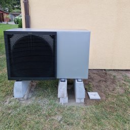 Eco green-Tech - Szara jednostka zewnętrzna urządzenia grzewczego Daikin, umieszczona na betonowych bloczkach na trawniku przy beżowej ścianie budynku, z widoczną rurą spustową.