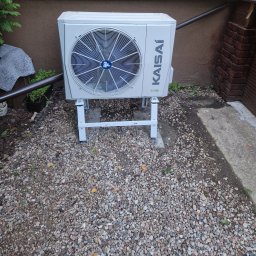 Eco green-Tech - Zewnętrzna jednostka klimatyzatora Kaisai na białym stelażu, ustawiona na żwirowym podłożu przy brązowej ścianie budynku i ceglanym narożniku.