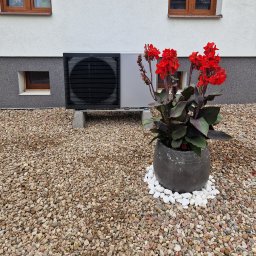 Eco green-Tech - Czarna zewnętrzna jednostka grzewcza na betonowych podstawach, obok duża donica z czerwonymi kwiatami na żwirowym podłożu przed budynkiem.