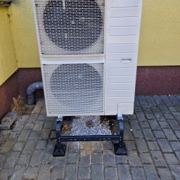 Eco green-Tech - Zewnętrzna jednostka dwuczęściowego systemu grzewczego, zamontowana na czarnej, metalowej podstawie z poziomującymi stopkami, umieszczona na szarej kostce brukowej, z szarą rurą odpływową po lewej...