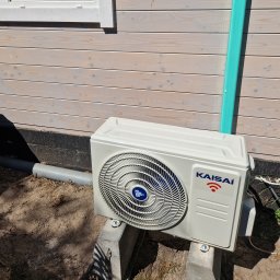 Eco green-Tech - Zewnętrzna jednostka klimatyzacji marki Kaisai zamontowana na betonowych blokach przy ścianie domu z jasnego drewna, widoczna rura odprowadzająca skropliny.