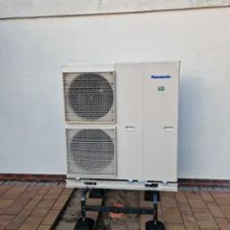 Zewnętrzna jednostka pompy ciepła Panasonic umieszczona na czarnym, metalowym stelażu na tle białej ściany i kostki brukowej.