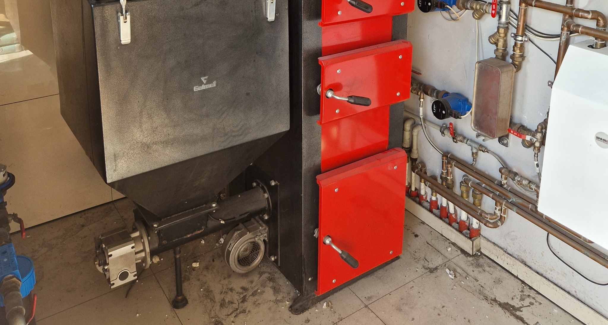 Częściowy widok kotła na paliwo stałe z czerwonymi drzwiczkami oraz instalacją hydrauliczną z rur miedzianych przy ścianie.