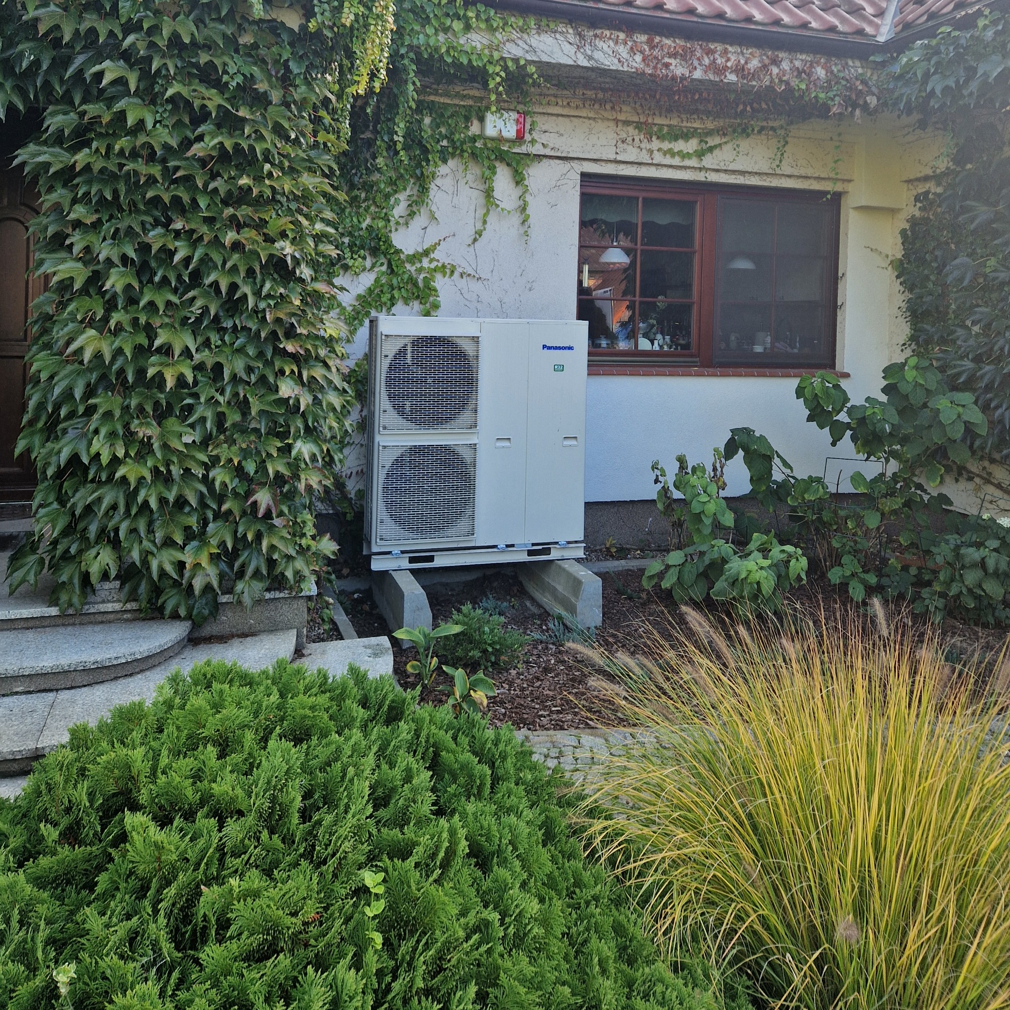 Biała jednostka zewnętrzna klimatyzacji Panasonic zamontowana na betonowych podstawach przed domem z oknem w brązowej ramie, elewacja częściowo porośnięta bluszczem.