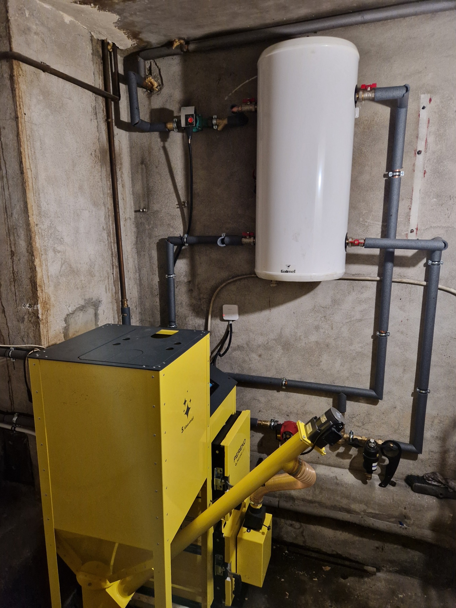 Żółty piec z podajnikiem, biały zbiornik na wodę i rury z izolacją w surowym pomieszczeniu z betonowymi ścianami.