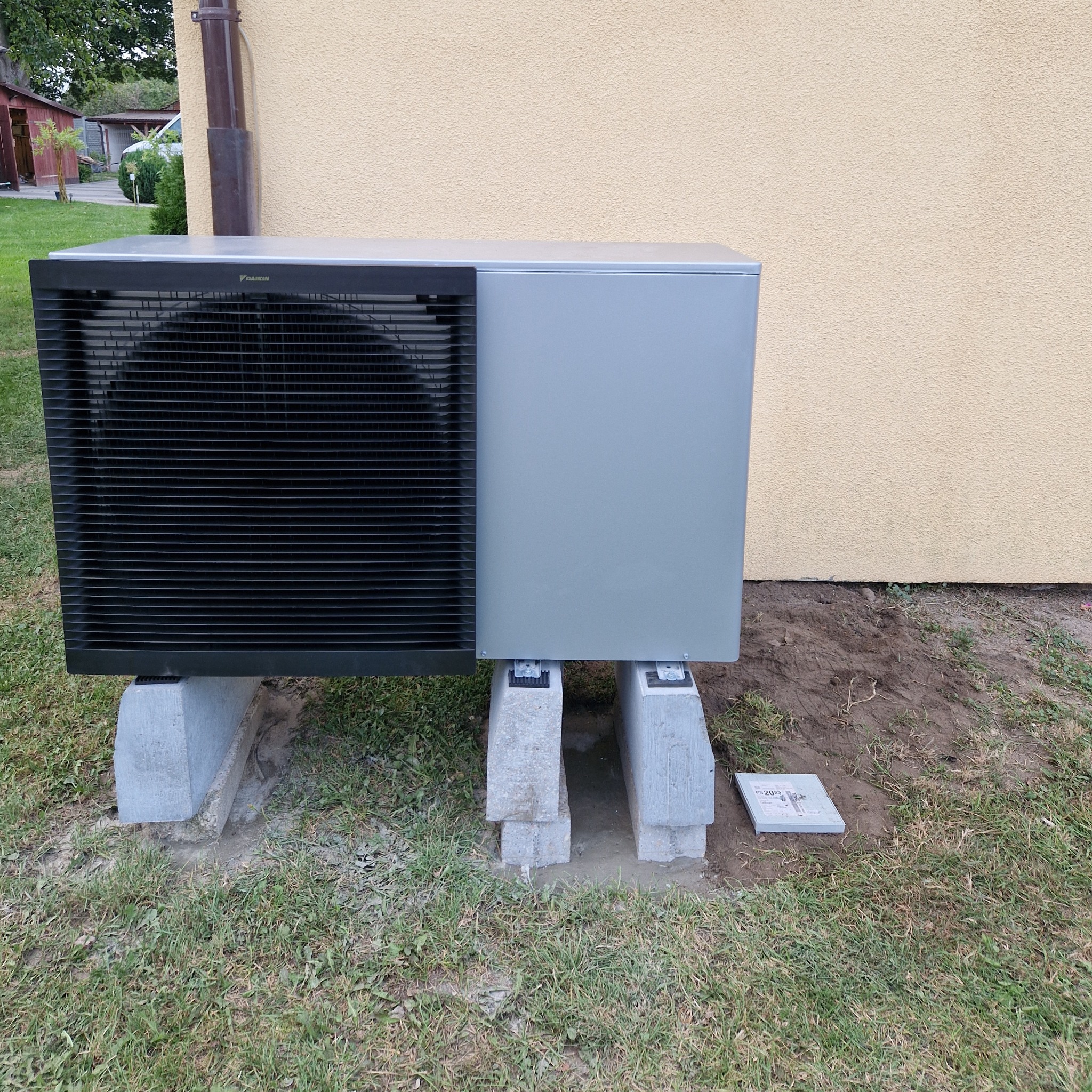 Szara jednostka zewnętrzna urządzenia grzewczego Daikin, umieszczona na betonowych bloczkach na trawniku przy beżowej ścianie budynku, z widoczną rurą spustową.