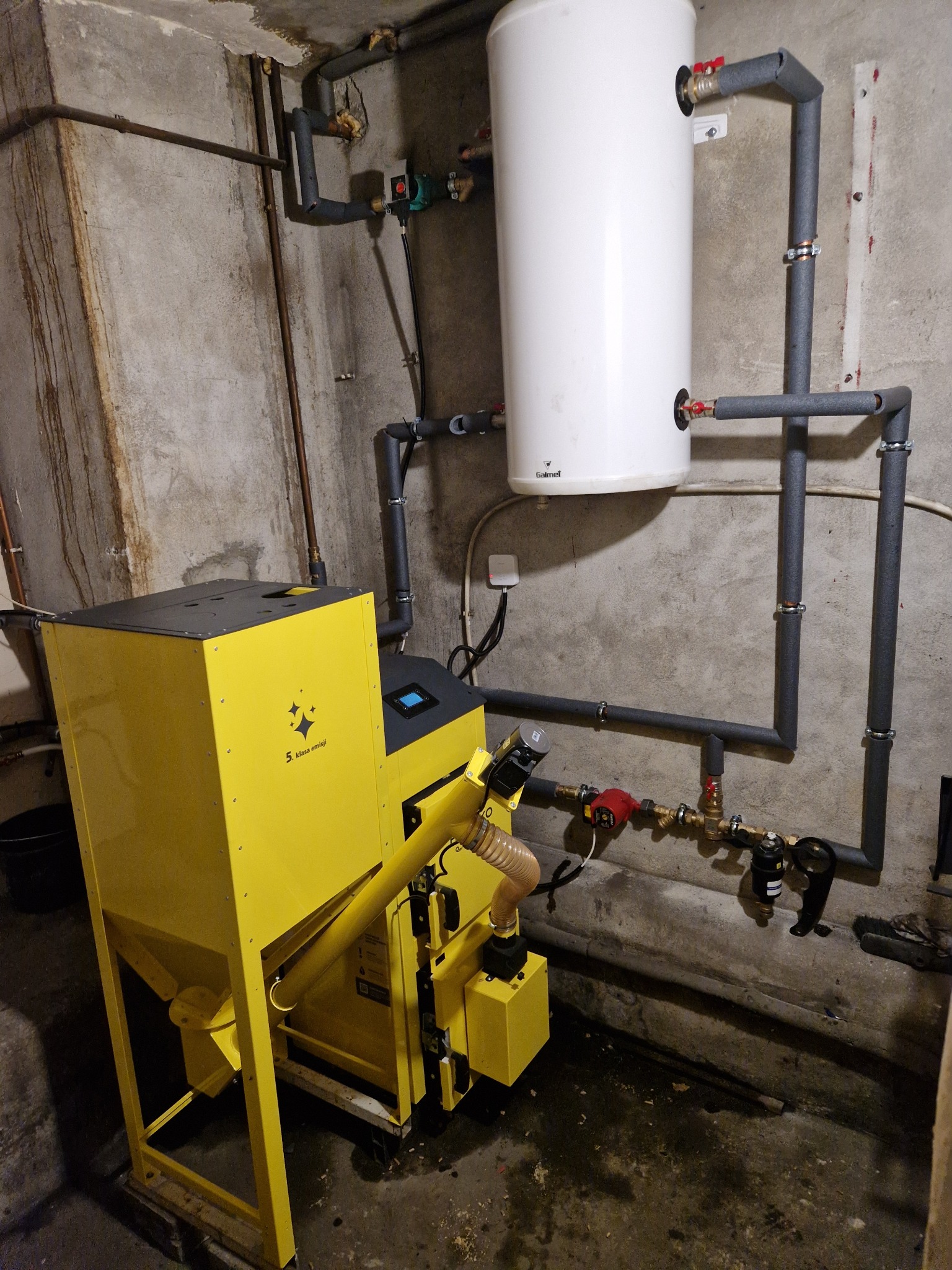 Żółty piec na pellet z podajnikiem ślimakowym, podłączony do instalacji grzewczej z białym zasobnikiem ciepłej wody użytkowej na tle surowej ściany kotłowni.