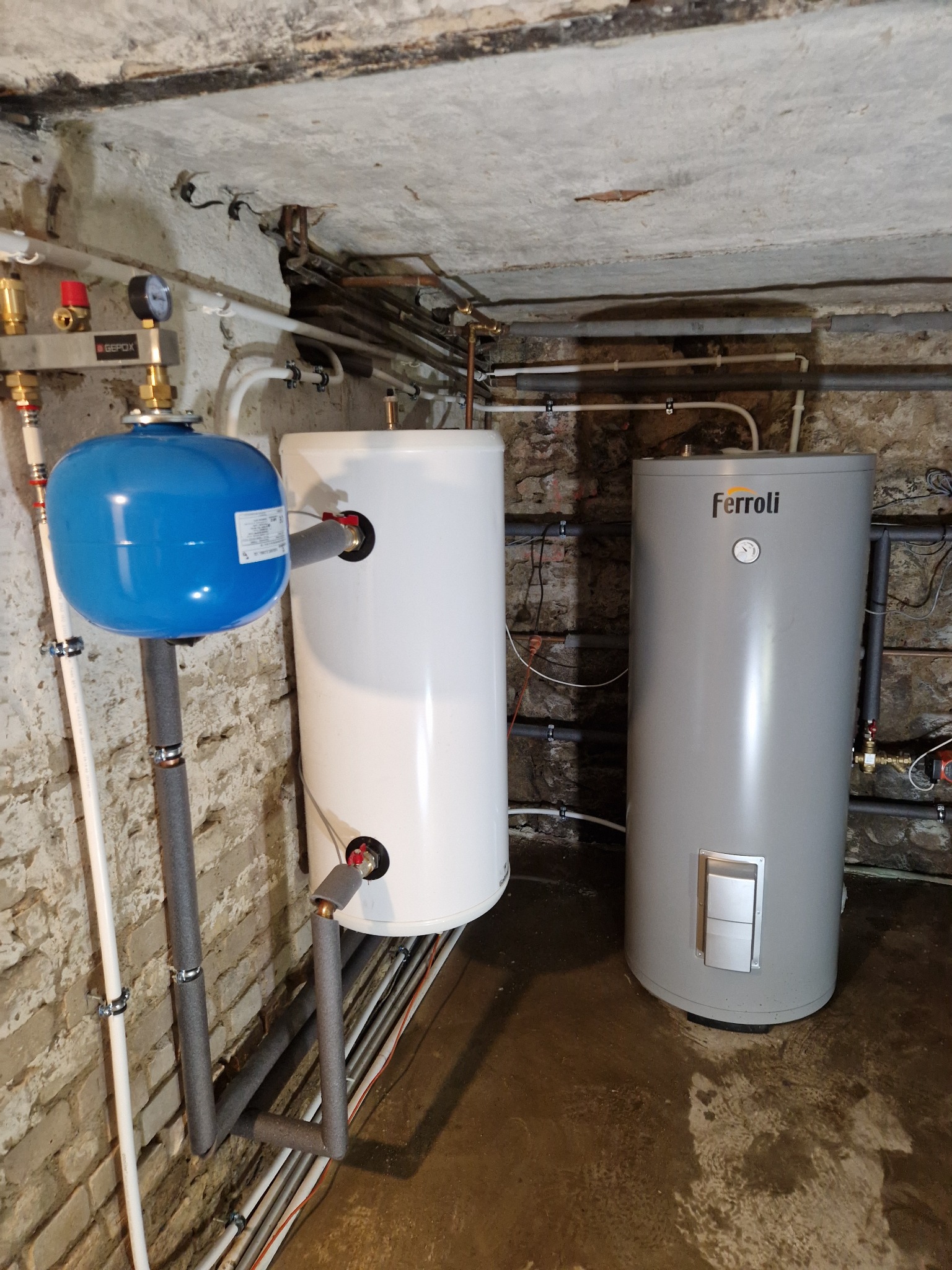 Instalacja zbiorników buforowych ciepłej wody użytkowej w piwnicy o surowych, ceglanych ścianach i betonowym suficie, z widocznym niebieskim naczyniem wzbiorczym i rurami w izolacji.