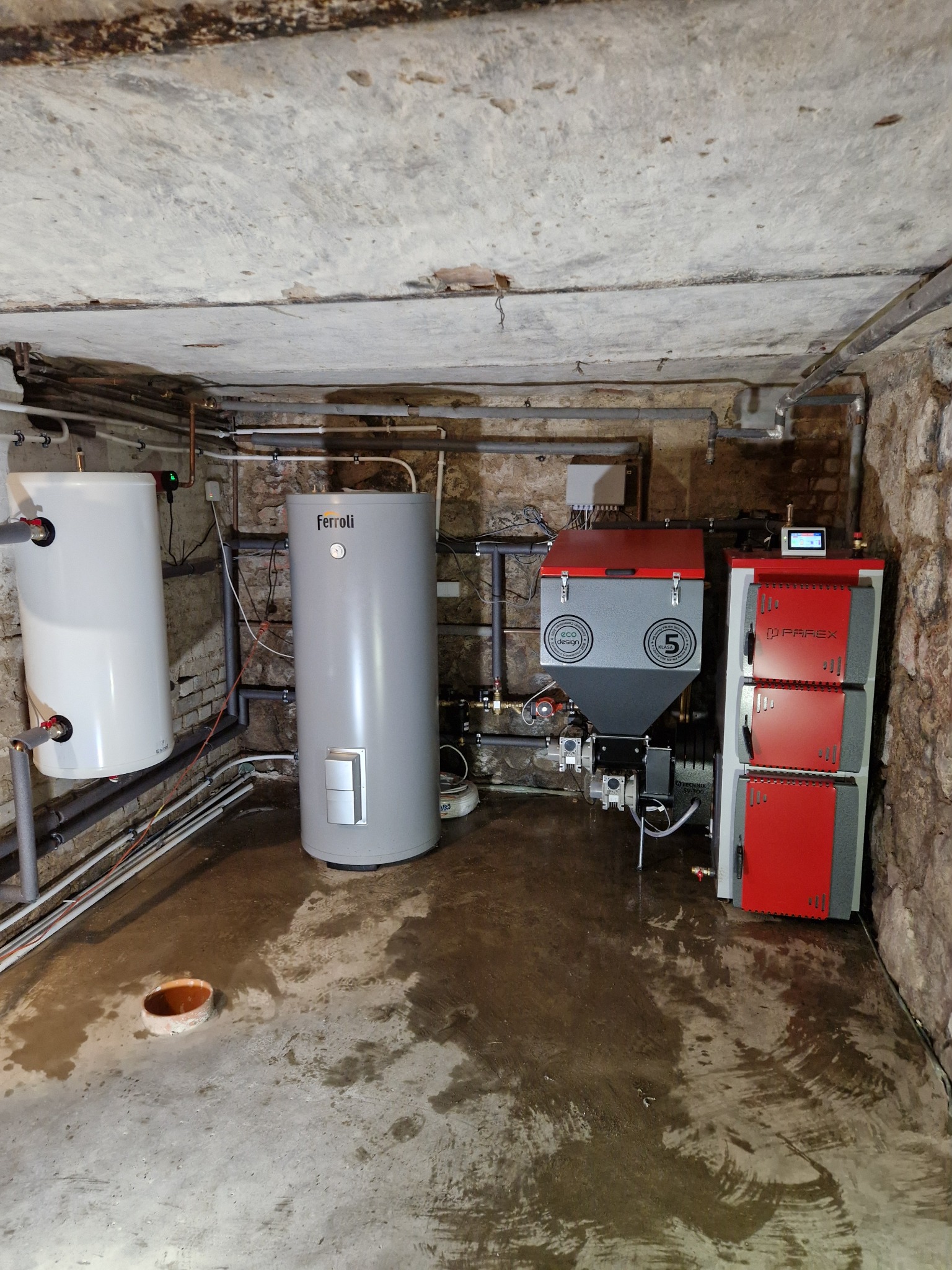 Piwnica z zainstalowanym kotłem na pellet z podajnikiem, dwoma zbiornikami wody, z widocznymi rurami instalacji grzewczej pod sufitem i wilgotną posadzką.